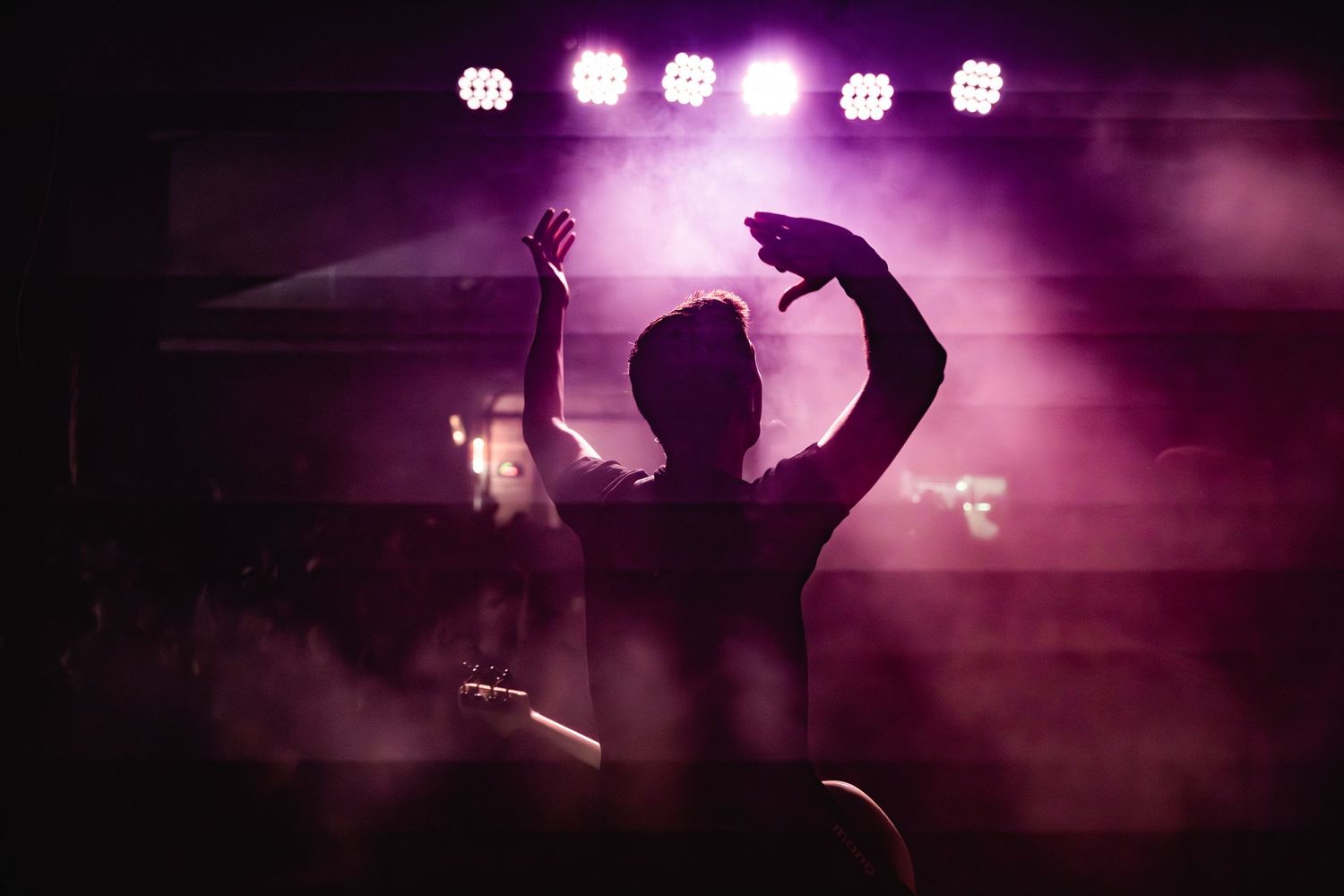 Silhouette of a concert attendee with raised arms against purple stage lights and fog.