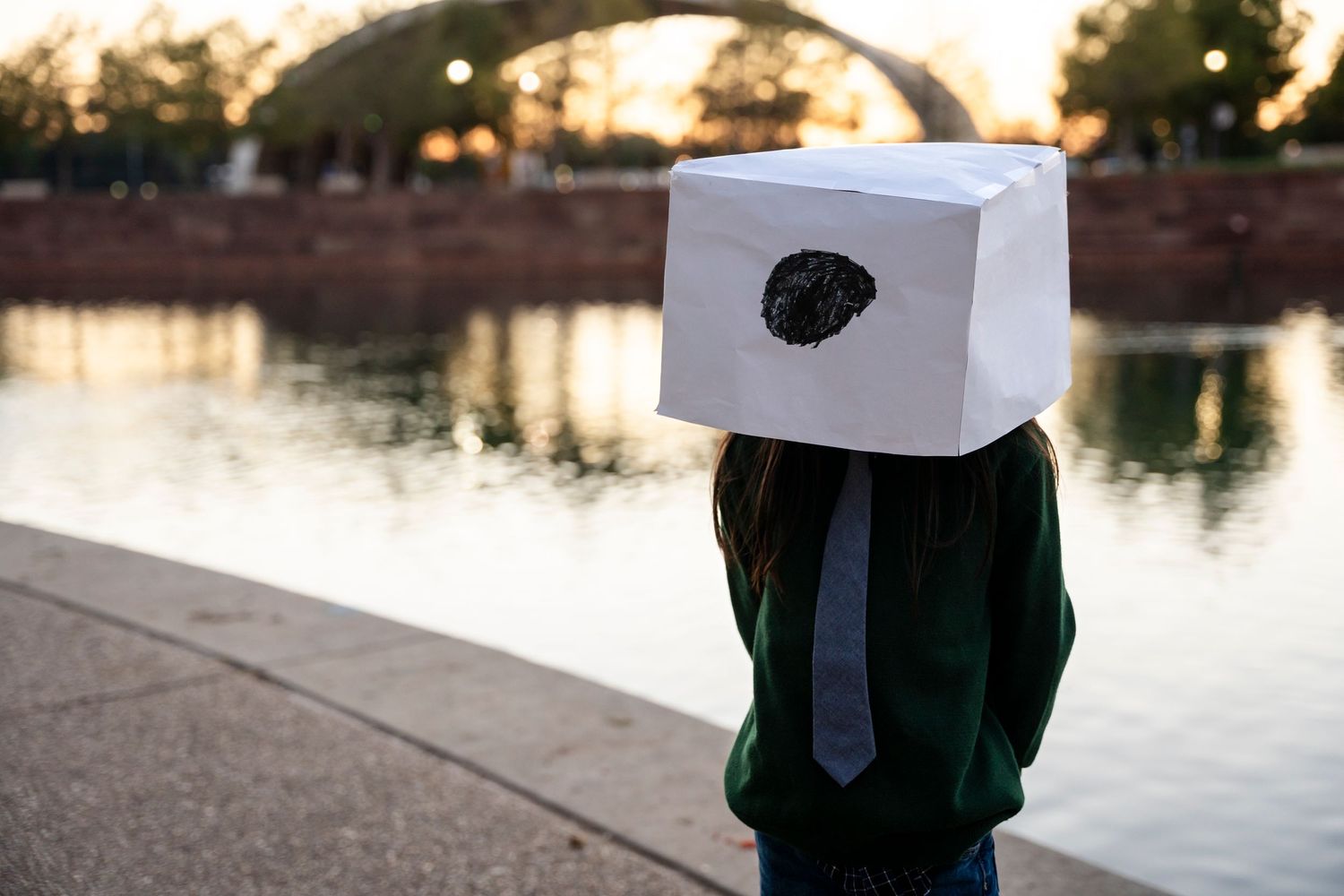 Person wearing green sweater with playful white box on head poses by lake.
