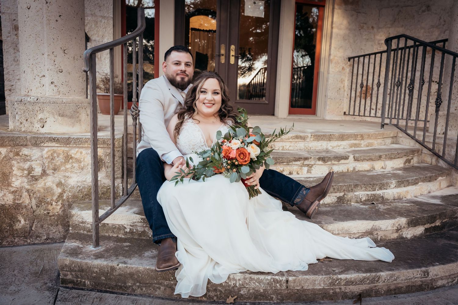 A couple sits on stone steps with the bride holding a vibrant orange and green bouquet.