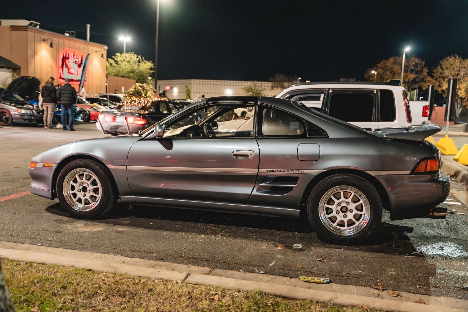 Modified grey Toyota MR2 parked on street curb at night.