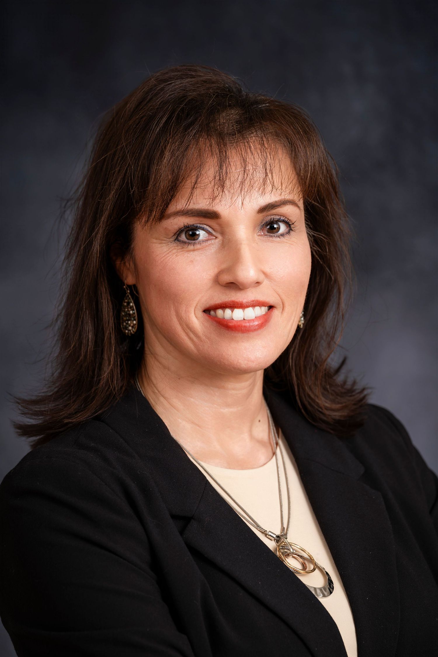 Professional portrait of a businesswoman wearing black blazer and jewelry against dark background.