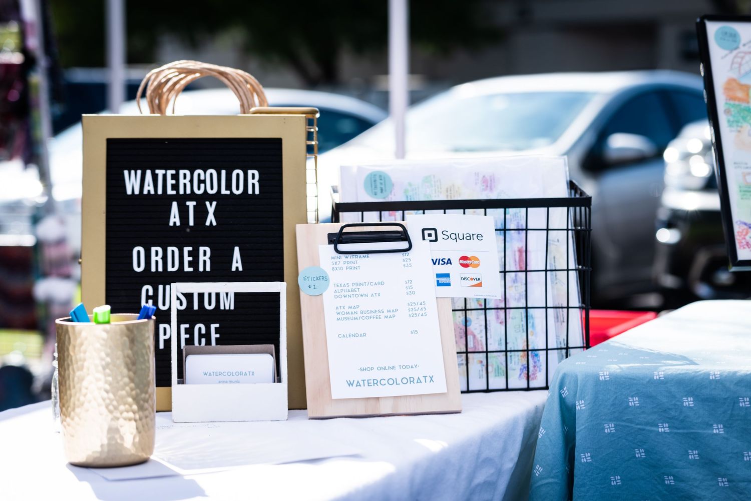 Sign advertising watercolor art and custom pieces at an outdoor market stall.