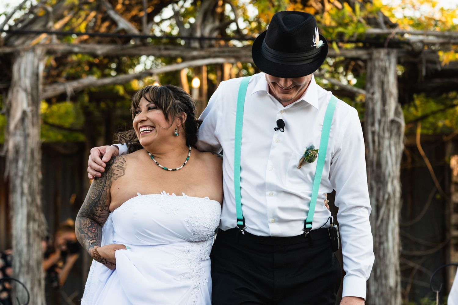 A couple shares a joyful moment under a wooden pergola at their outdoor wedding.