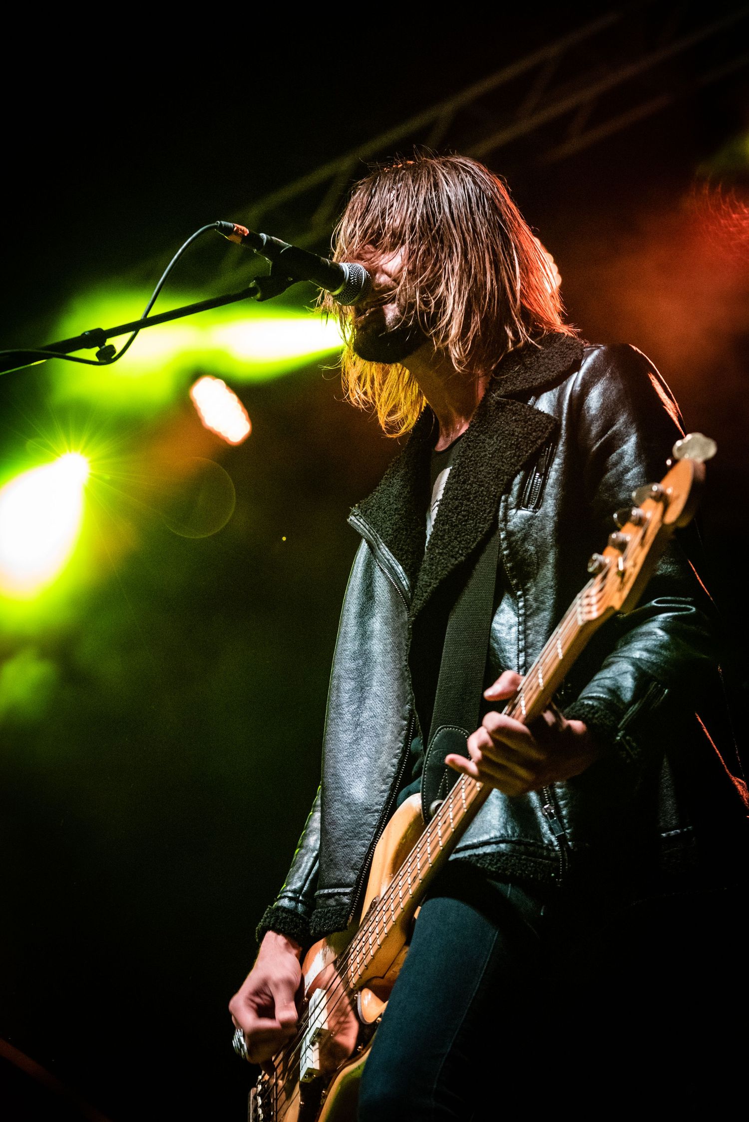 Rock musician with long hair performs on stage with bass guitar under dramatic green and orange lighting.
