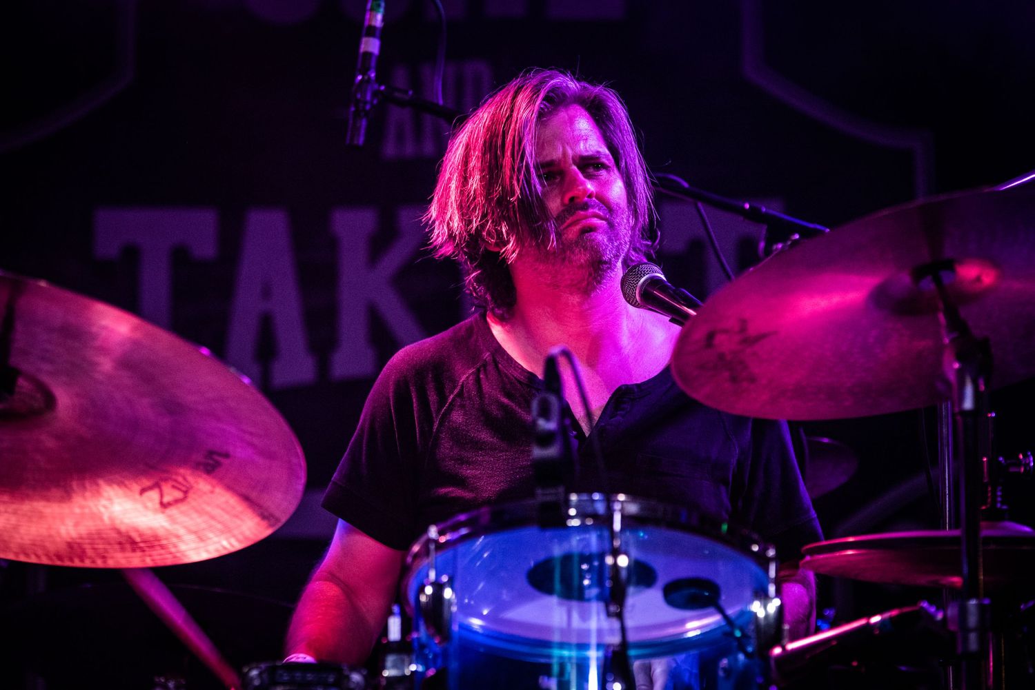 Drummer performing on stage bathed in purple lighting during live concert.