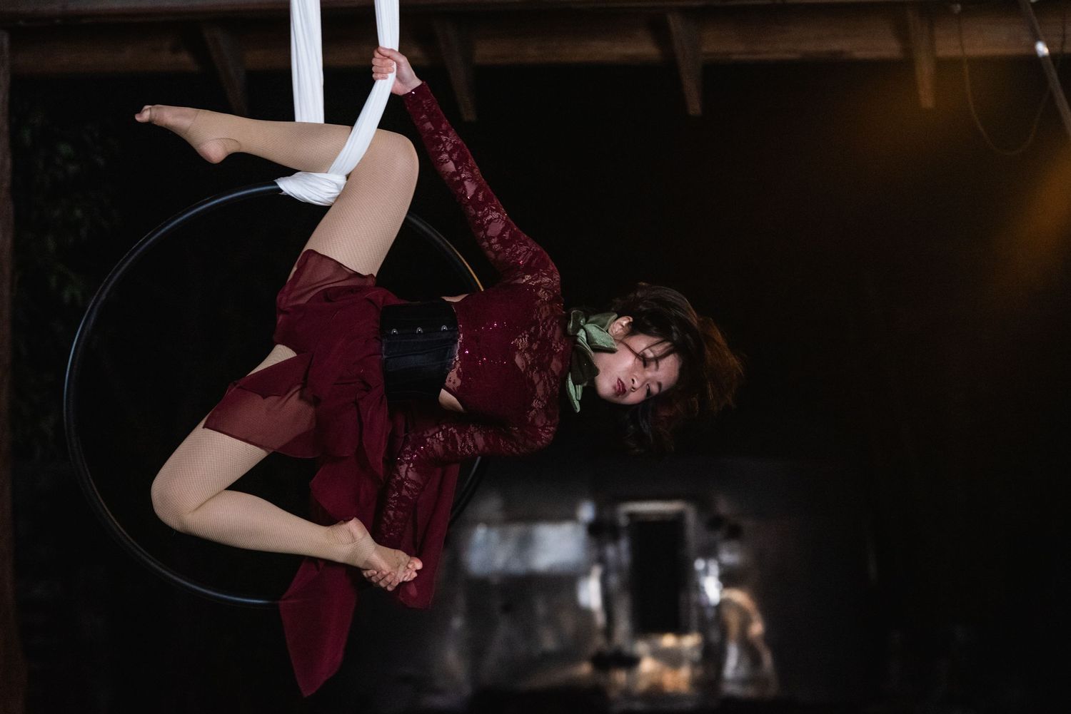 Aerial performer in burgundy dress gracefully poses on lyra hoop against dark background.