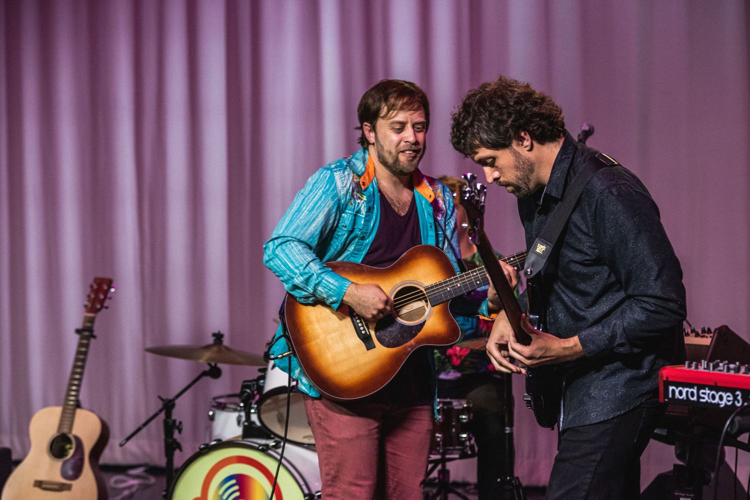 Two musicians perform together with acoustic and electric guitars on stage with purple backdrop.