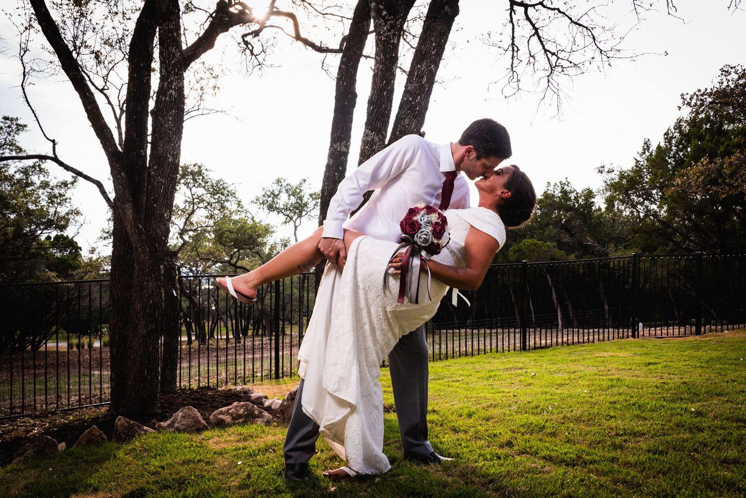 Newlyweds share romantic dip kiss on grassy lawn with iron fence and trees in background.