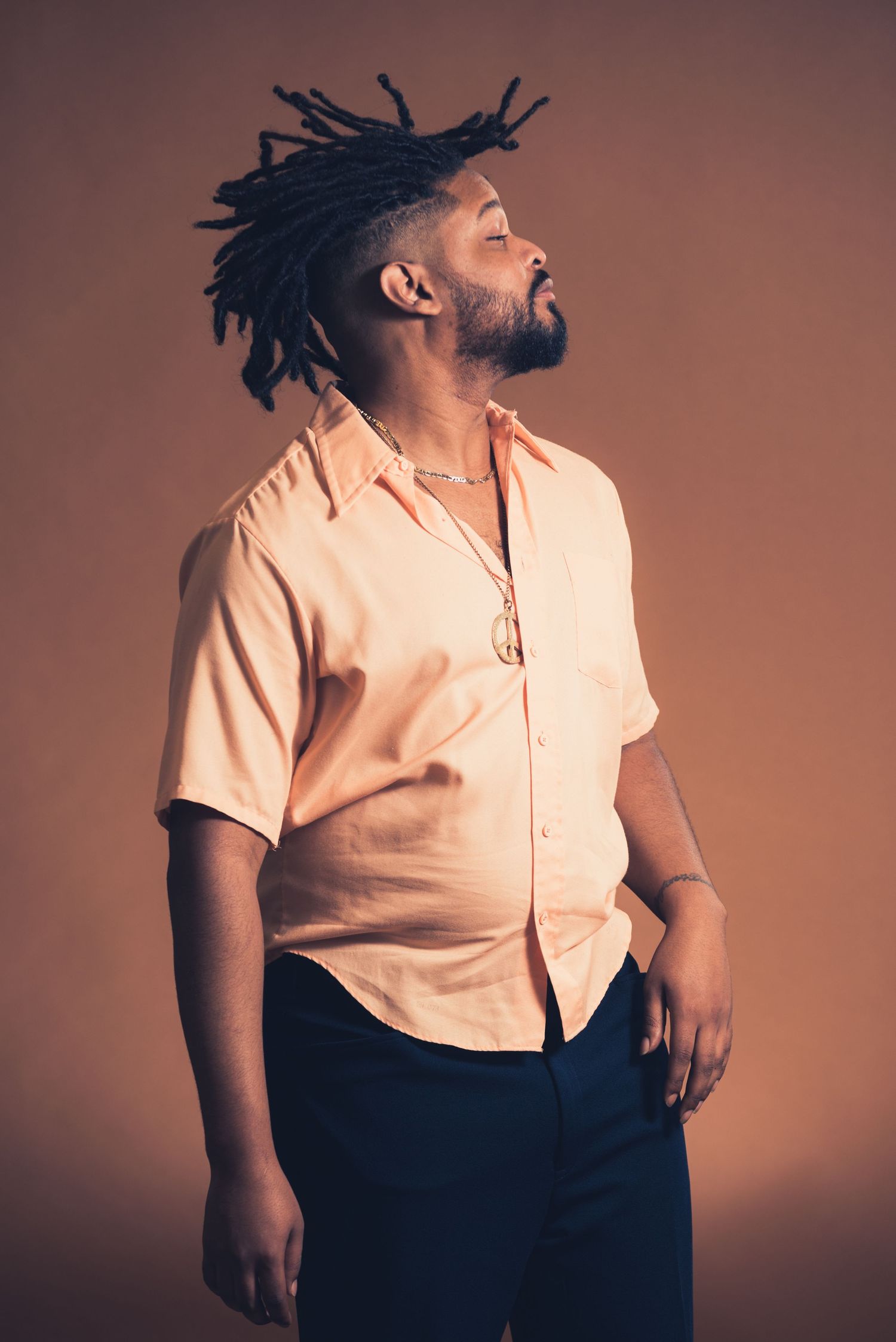 Portrait of person in peach button-up shirt with dreadlocks looking upward against brown background.