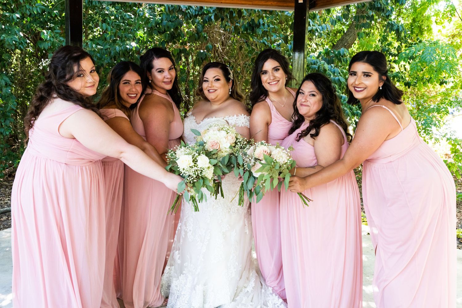 Bridesmaids in pink dresses embrace bride in white gown under outdoor gazebo.