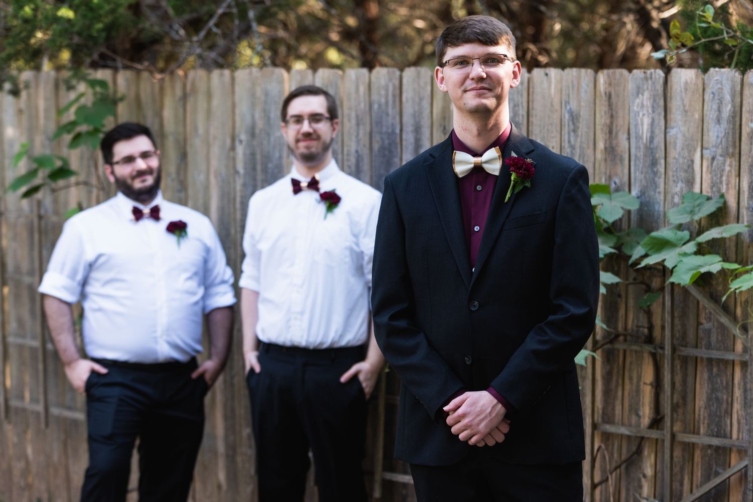 Three groomsmen stand in formal attire against a wooden fence backdrop at a wedding.