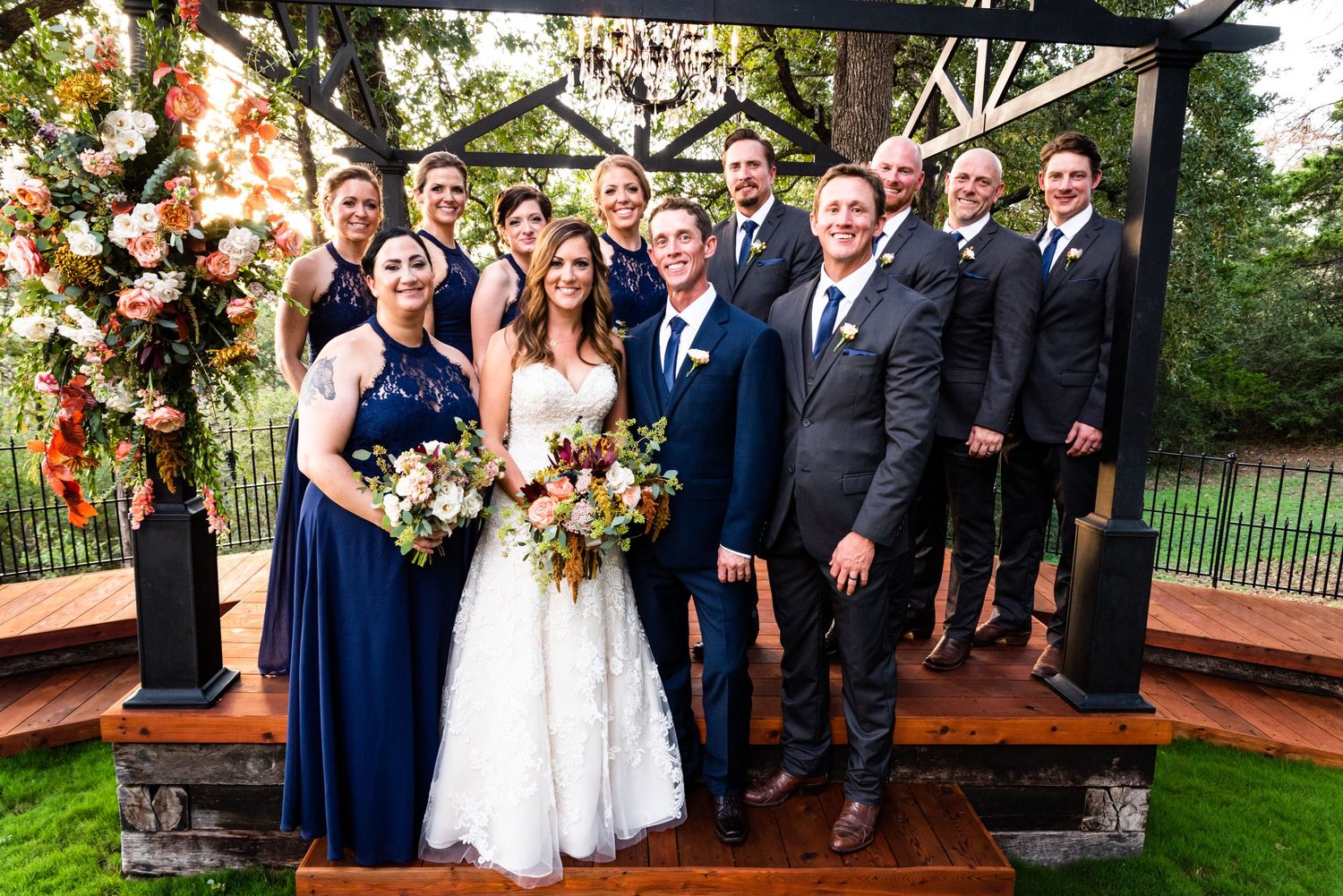 A wedding party poses together on a wooden deck with floral decorations and navy blue attire.
