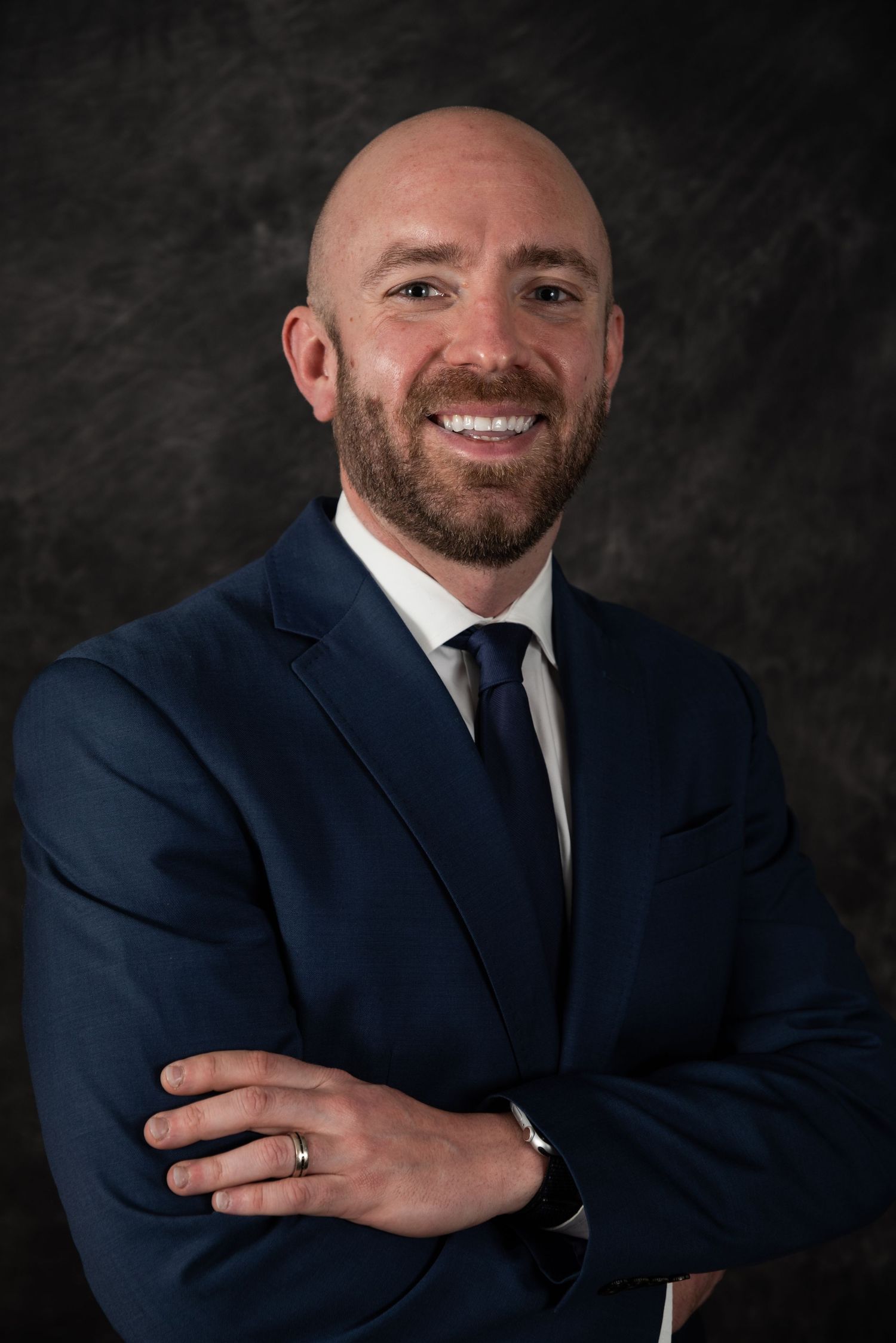 Business professional poses in navy suit for corporate headshot against dark background.