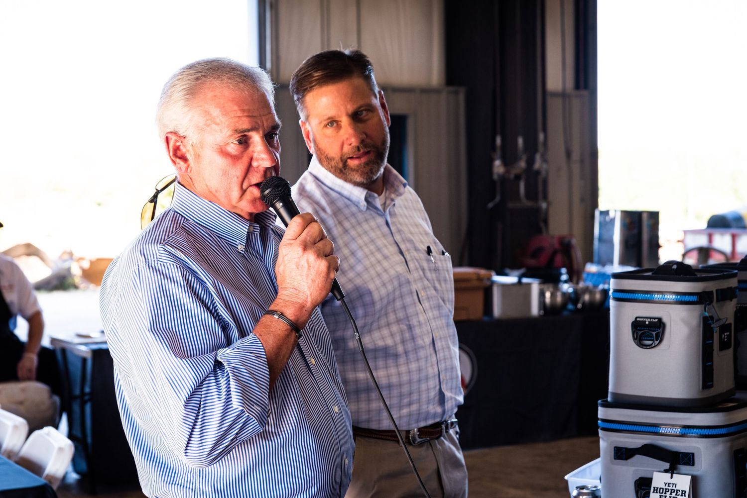 Two people speaking at an indoor event with coolers visible in background.