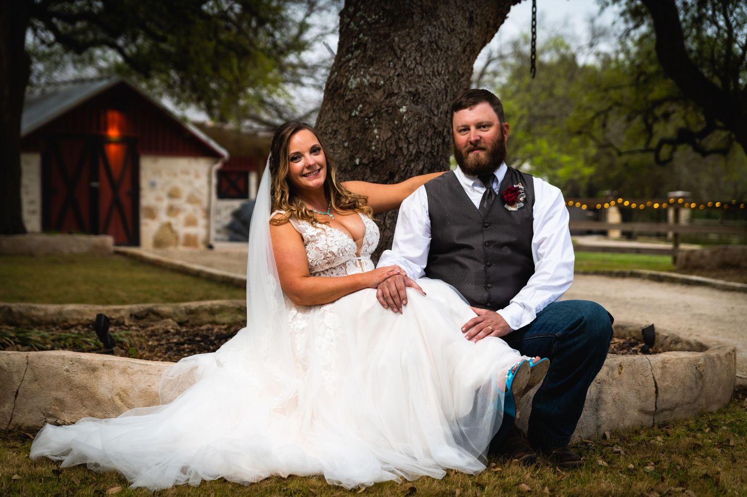 A bride in a white gown poses with her groom wearing a vest while sitting outdoors near a rustic building.