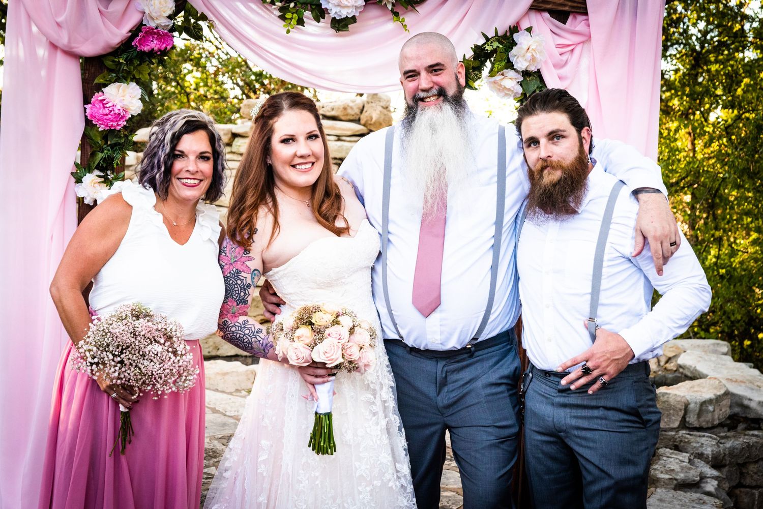 Wedding party members in pink dresses and blue suits posing under floral arch.