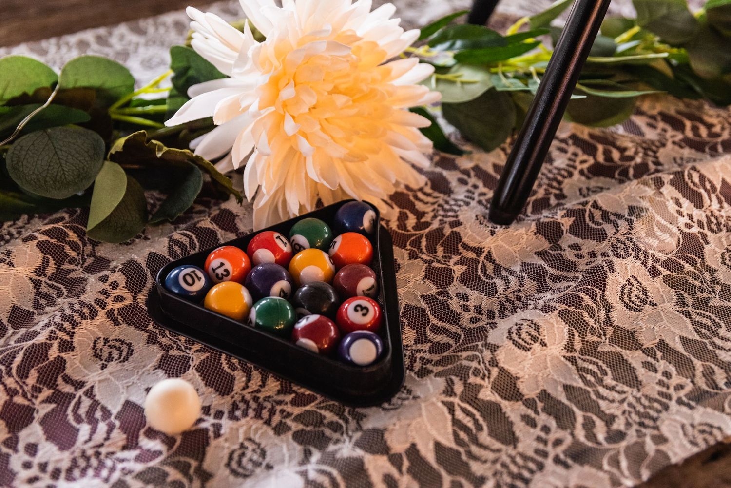 A peach chrysanthemum flower beside a triangular rack of pool balls on patterned lace fabric.