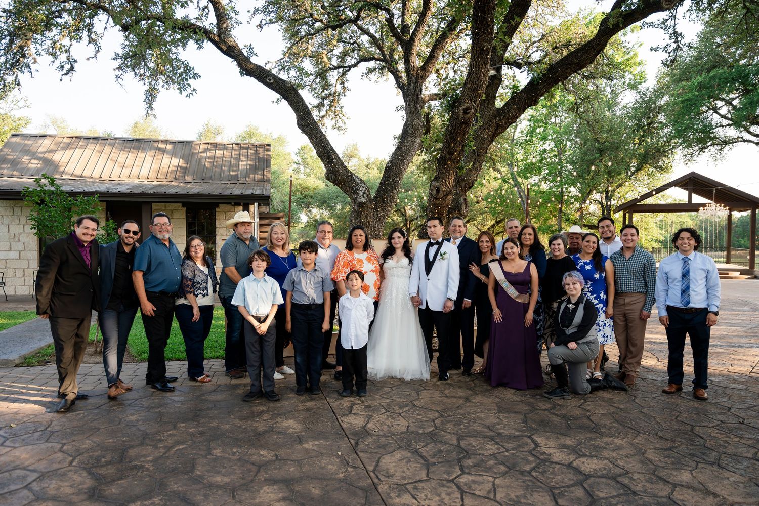 Wedding party and guests gathered for a group photo outdoors at a rustic venue with stone walls and overhanging trees.