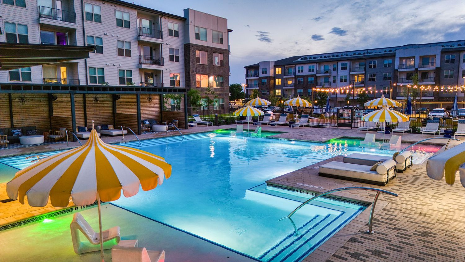 Evening view of lit swimming pool with yellow striped umbrellas at luxury apartment complex.