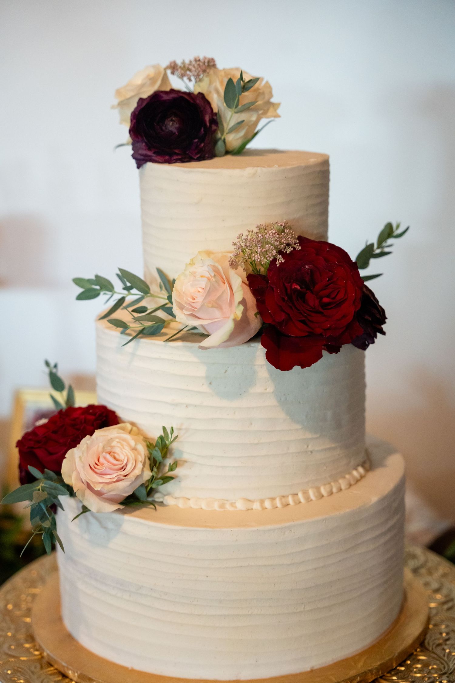 Three-tiered white wedding cake adorned with burgundy and blush roses and eucalyptus leaves.