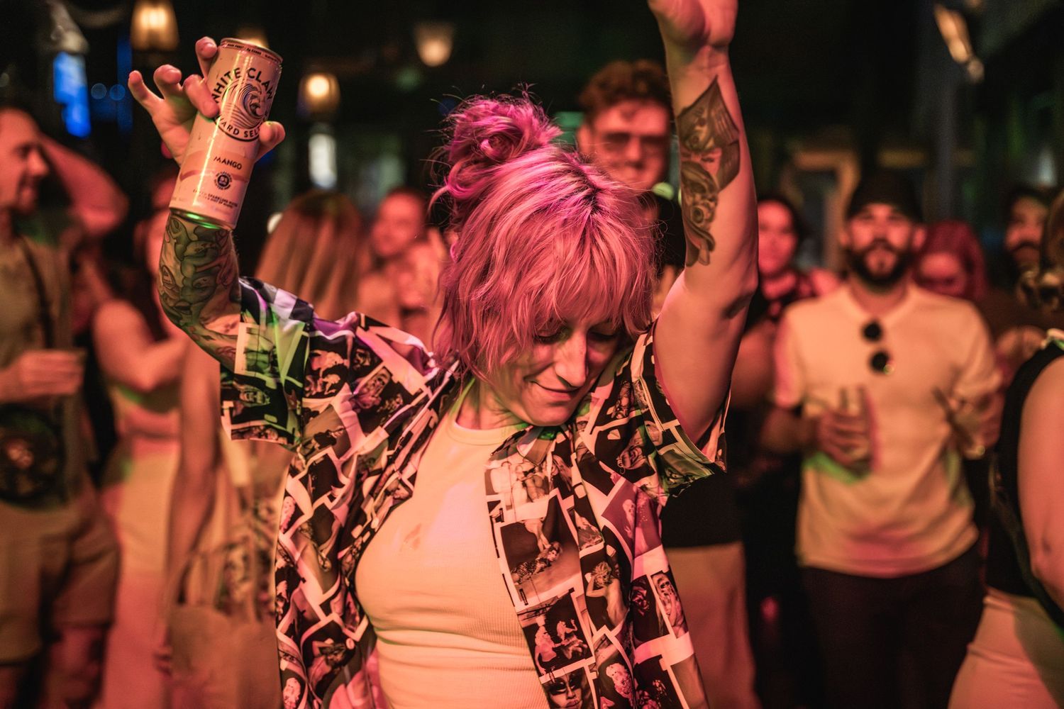 Concert-goer with pink hair dances with raised arms in a patterned shirt among the crowd.
