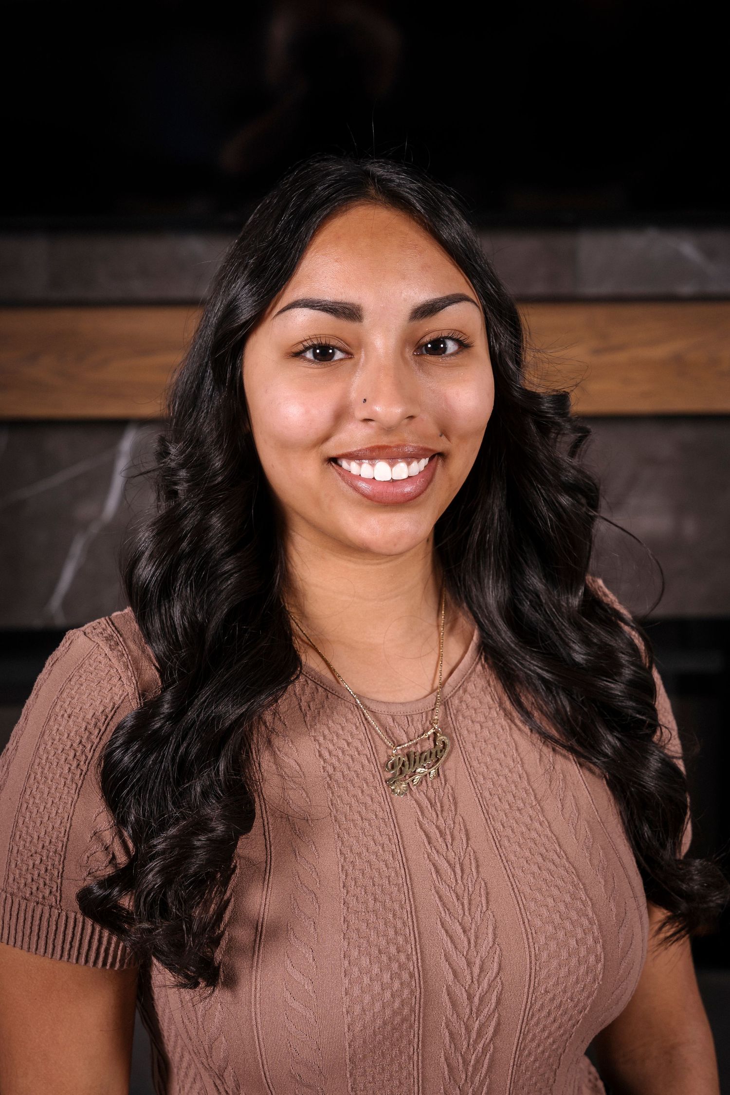 Portrait of a young woman with long dark wavy hair wearing a brown cable knit sweater and necklace against dark background.