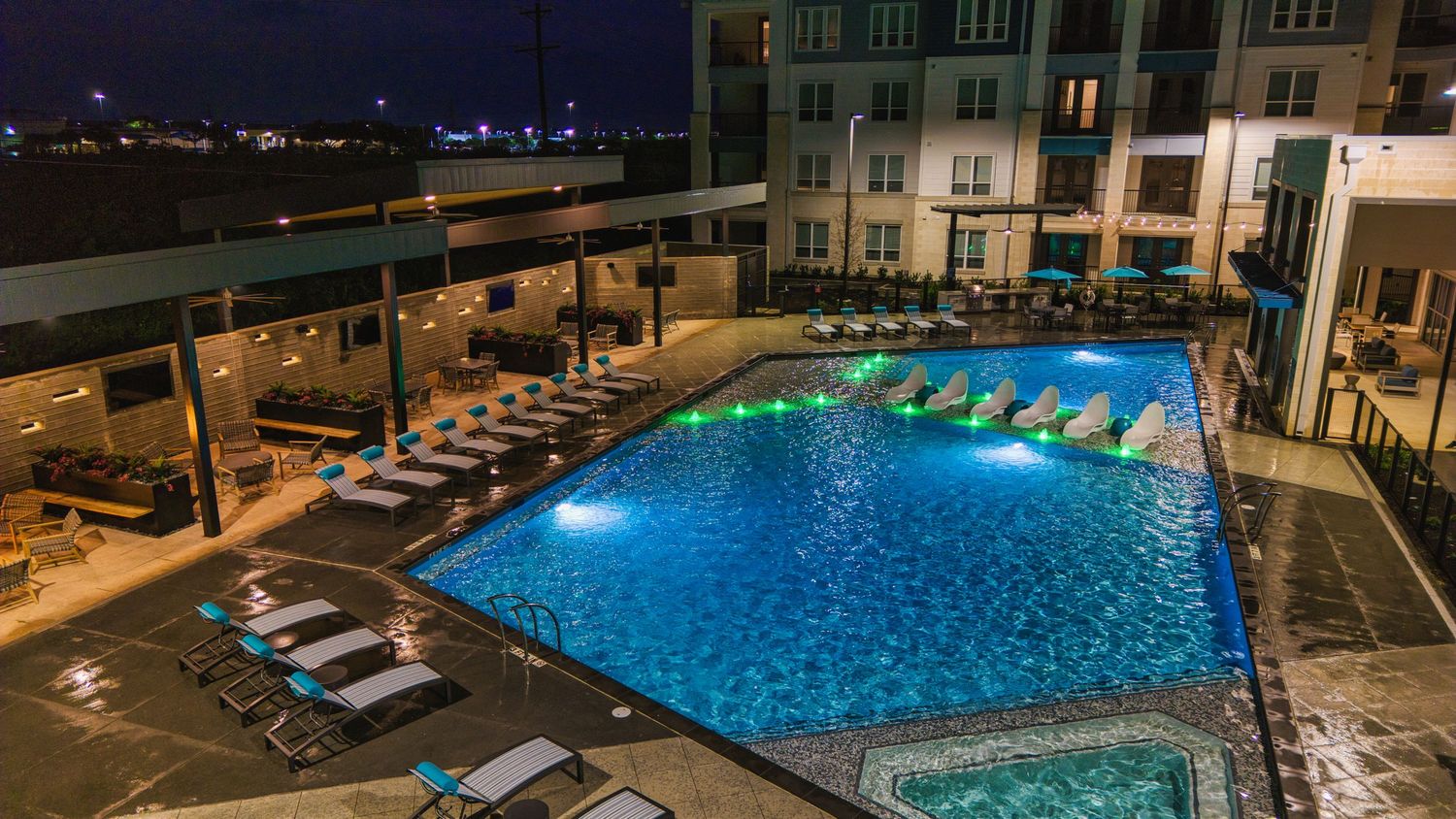 Illuminated apartment complex pool with blue LED lighting and lounge chairs at night.