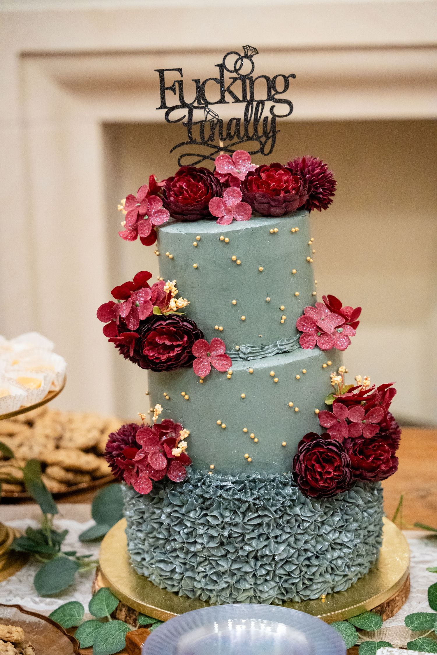Three-tiered blue wedding cake decorated with burgundy flowers and pearl details.
