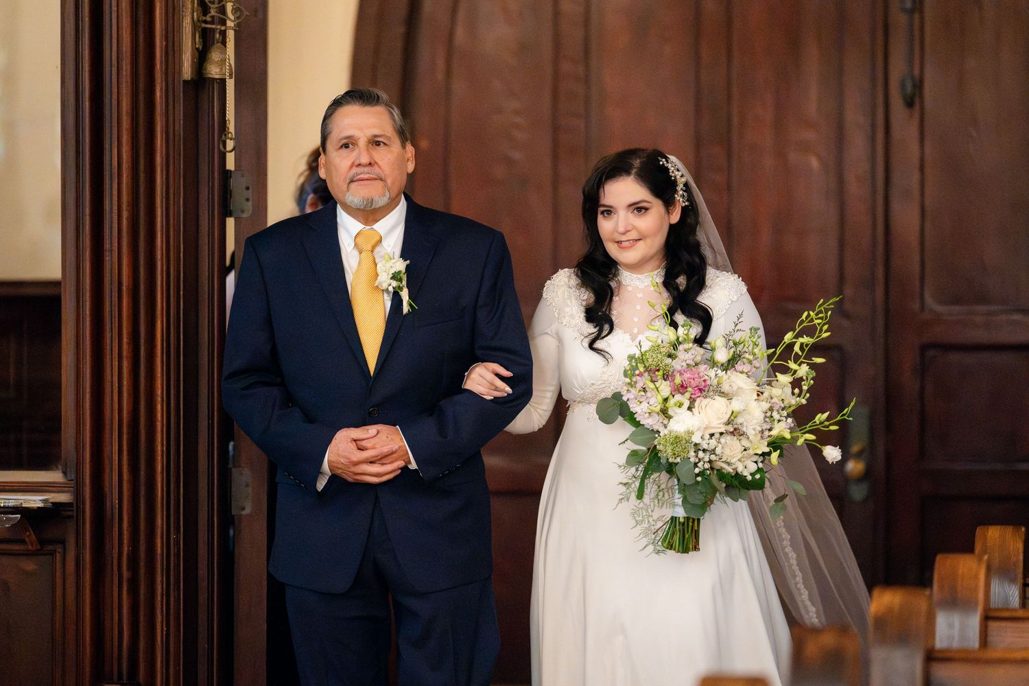 A bride in a white wedding dress holds a bouquet while standing by someone in a navy suit with yellow tie in a church.