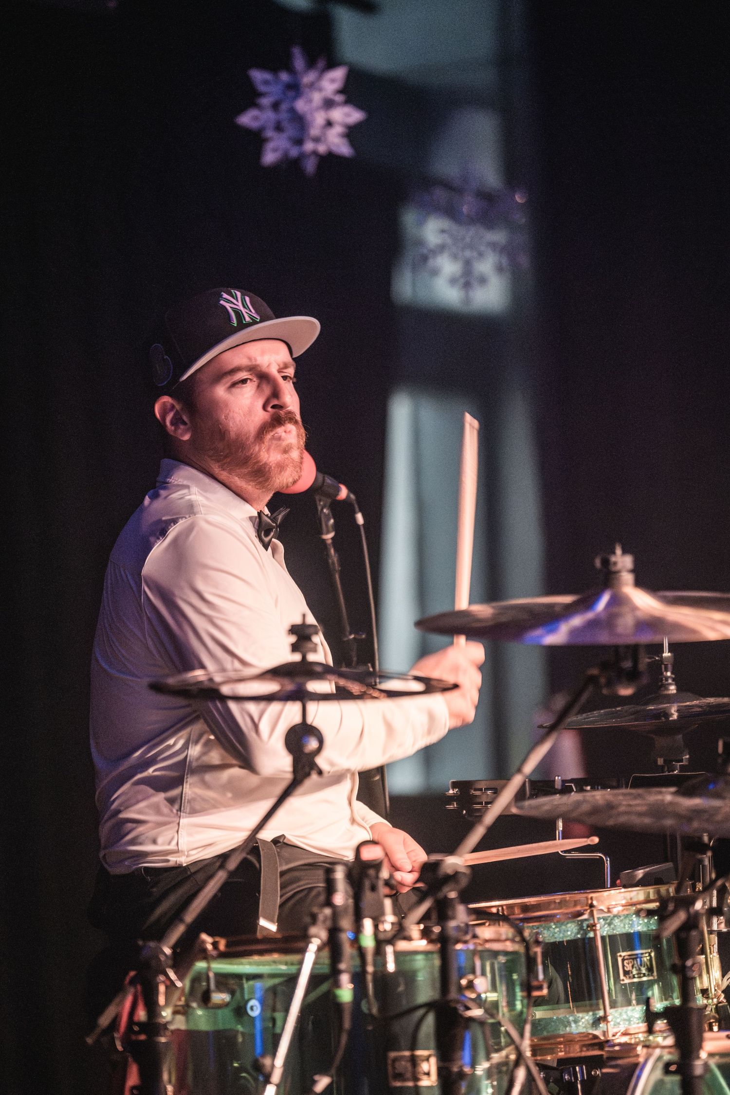Drummer wearing baseball cap performs on stage with purple lighting and star decoration.