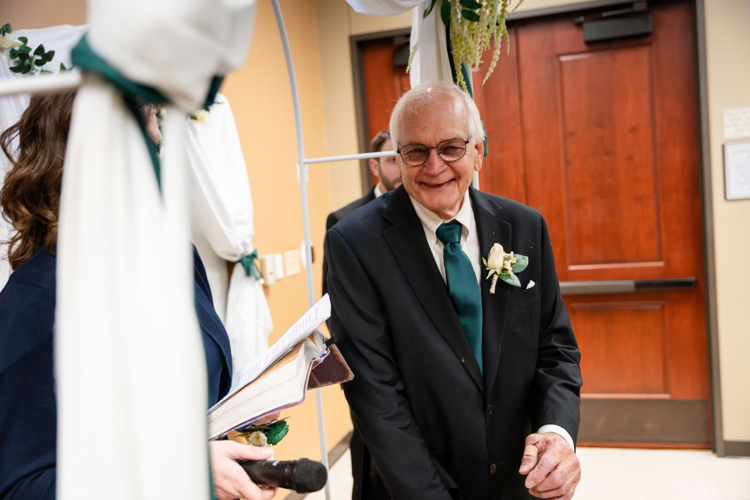 An elderly gentleman in a black suit and teal tie smiles warmly while wearing a boutonniere at a wedding celebration.