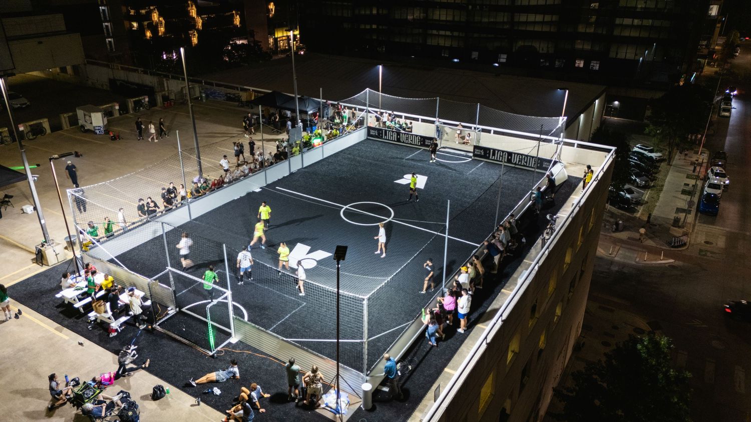 Aerial view of a rooftop soccer court lit up at night with spectators watching the game.