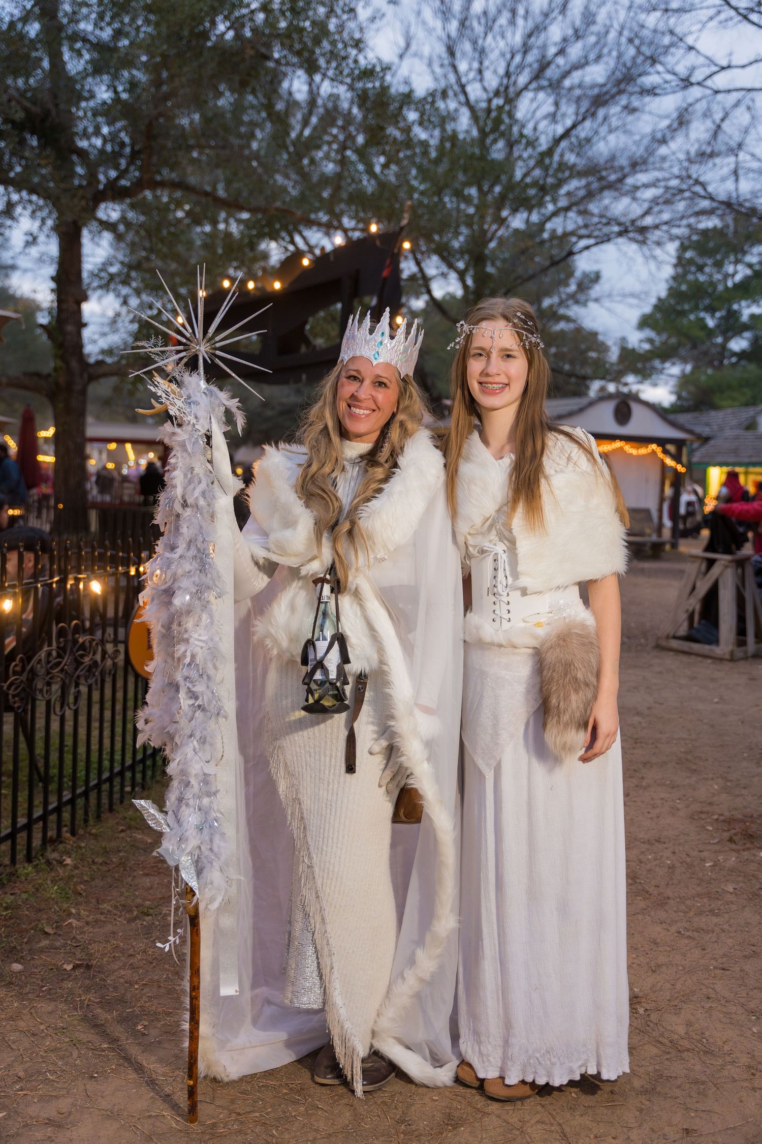 Two performers in ethereal white winter queen costumes hold decorative staffs at twilight.