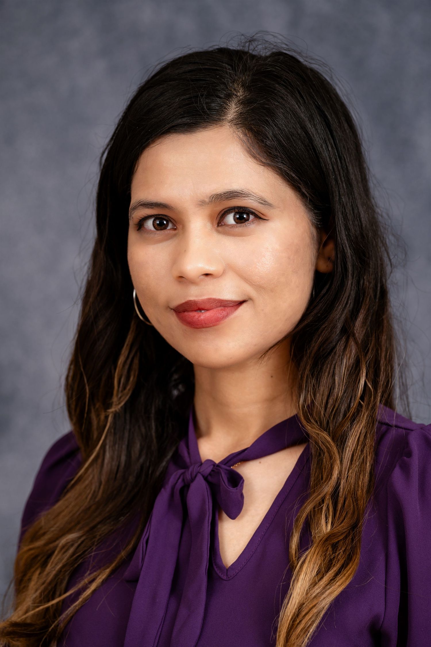 Professional headshot of a person in a purple tie-neck blouse against a gray background.