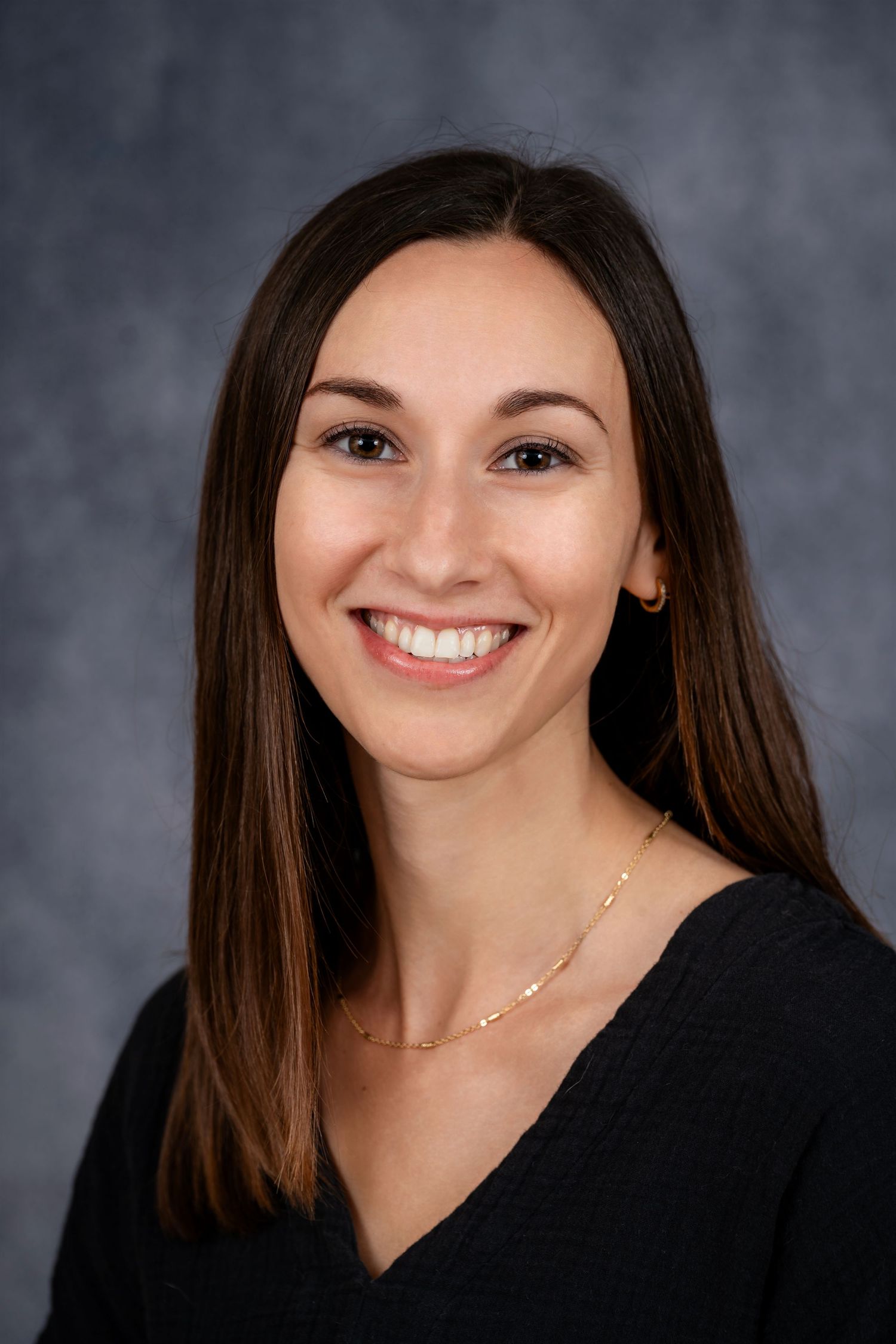Professional portrait in black v-neck top with delicate gold necklace against gray background.