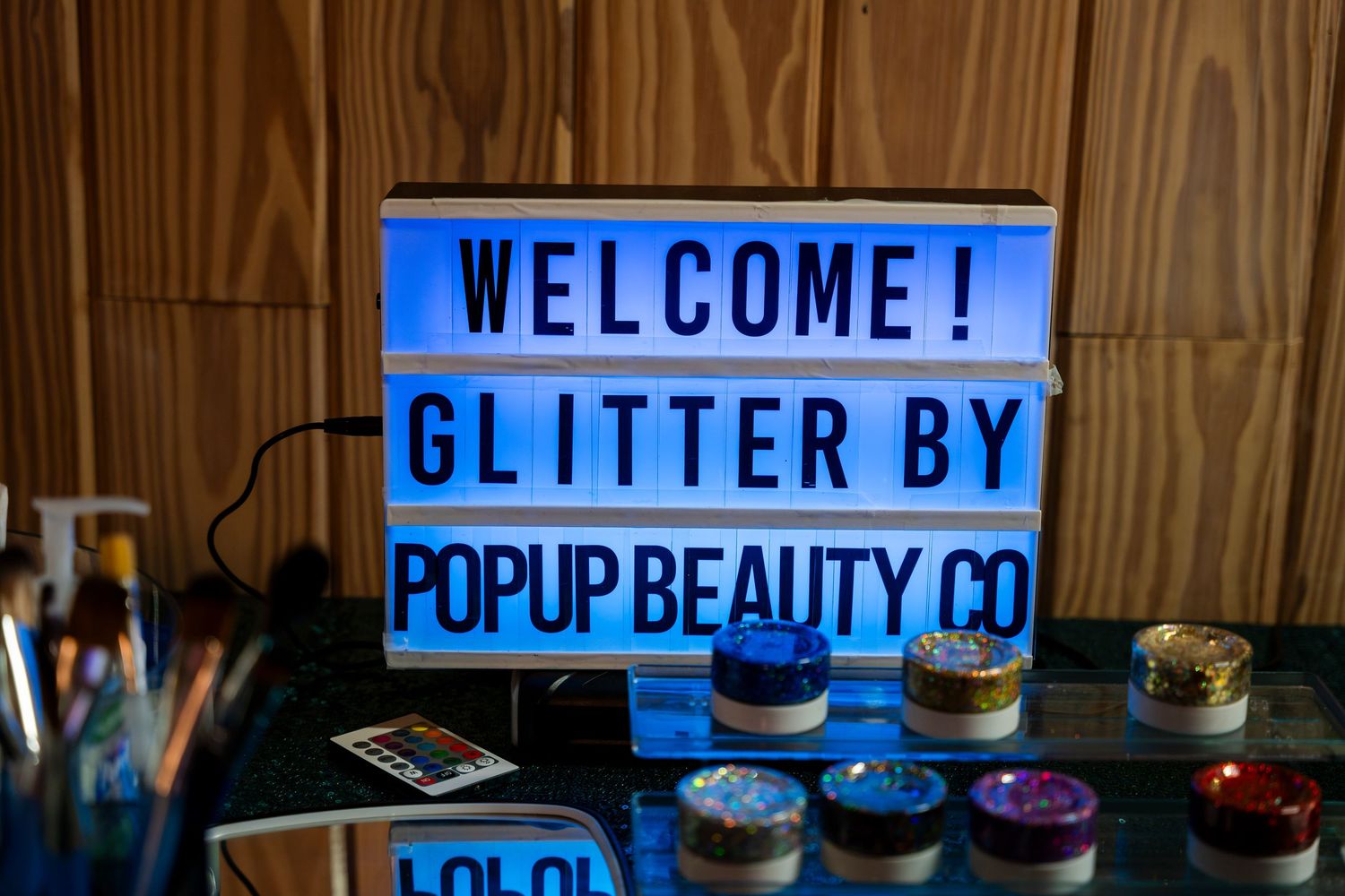 An illuminated welcome sign for Glitter by PopUp Beauty Co with colorful glitter containers displayed on a wooden table.