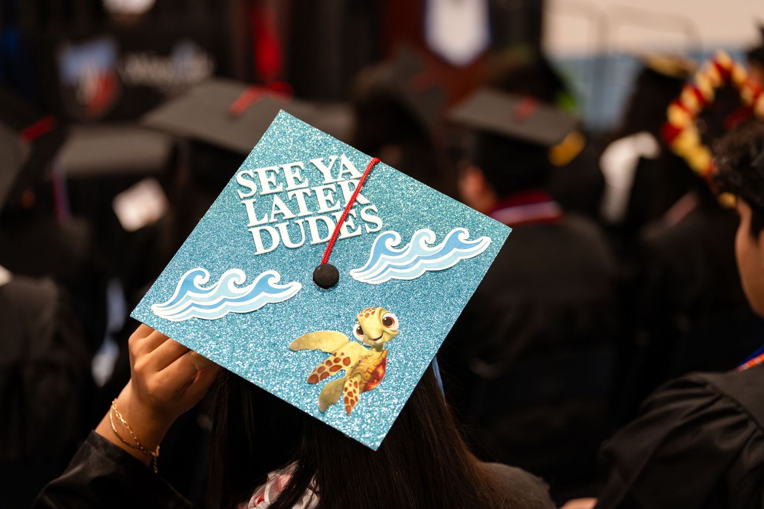A decorated graduation cap with a surfing theme featuring waves and text 'Seas the day dude' in turquoise blue.