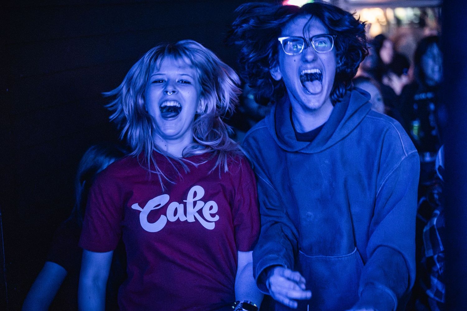 Two excited people at a concert or event, one wearing a red Coca-Cola shirt and another in a blue hoodie, reacting with joy.