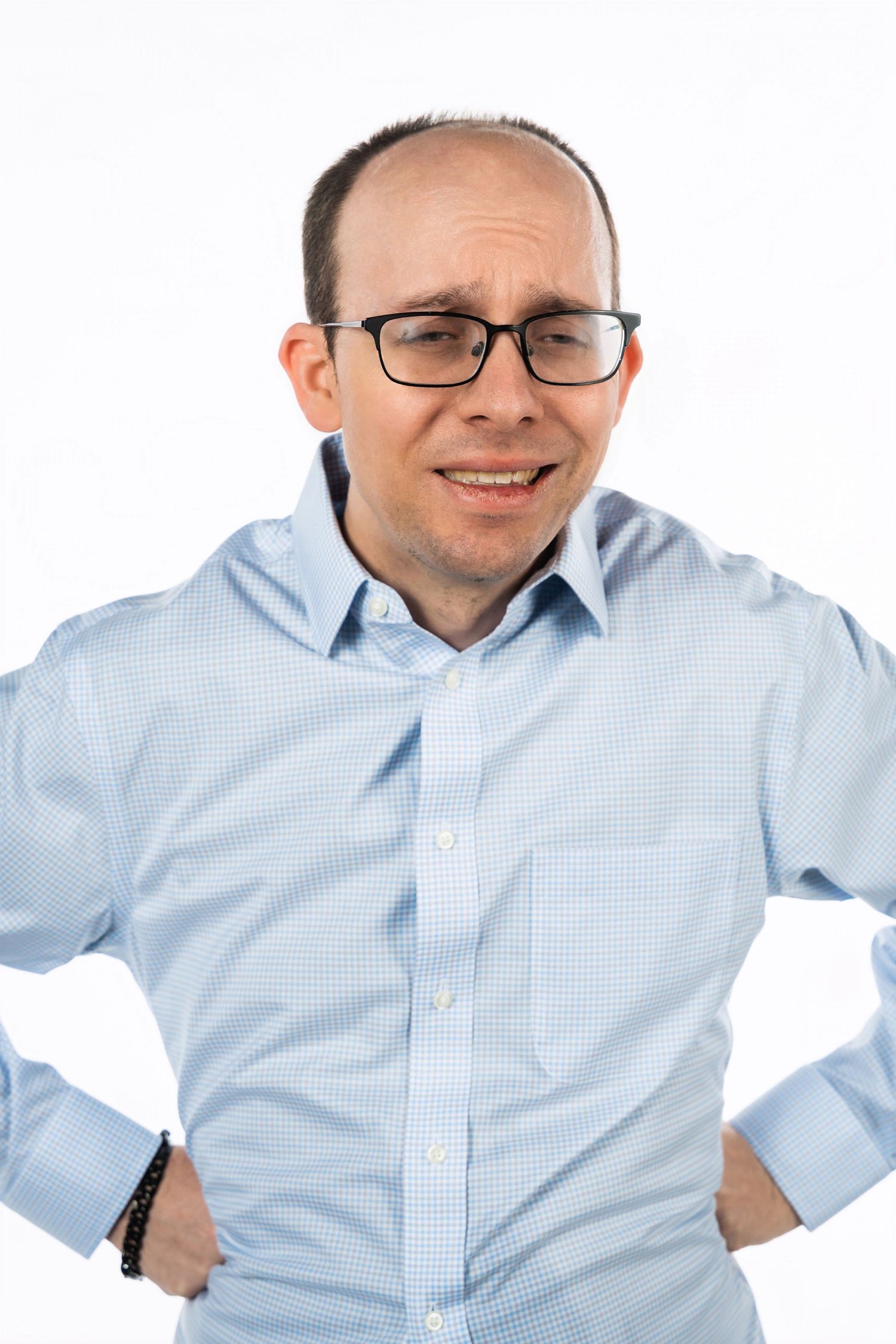 A person wearing glasses and a light blue dress shirt poses with hands on hips against white background.