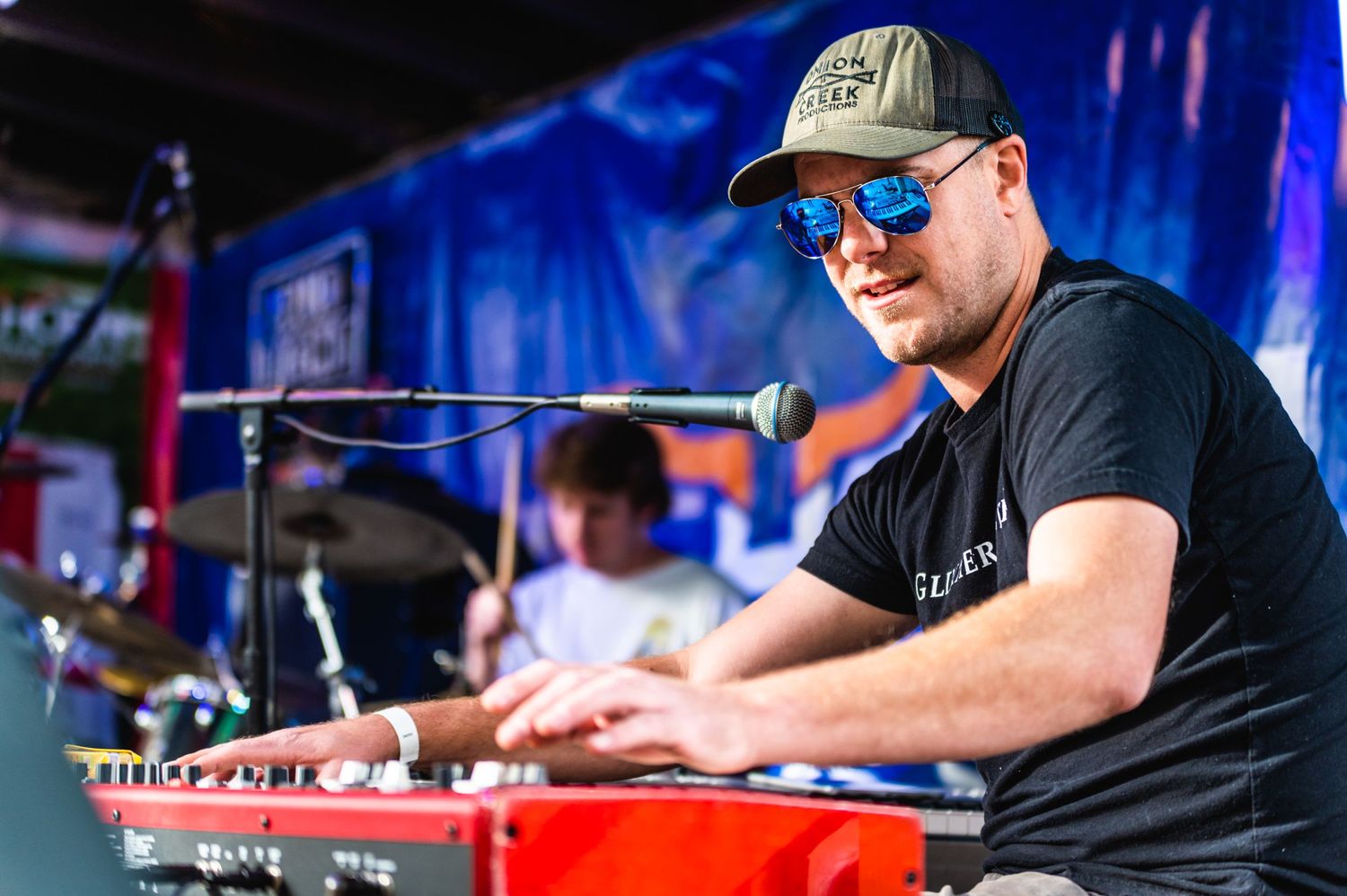 Keyboard player in black t-shirt and cap performs on red keyboard at outdoor concert.