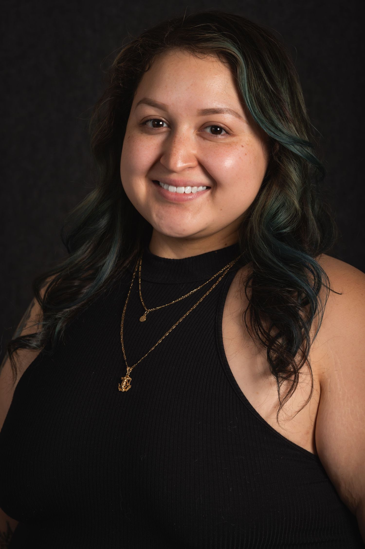 Portrait of a person wearing a black top and layered necklaces with teal-tinted hair.