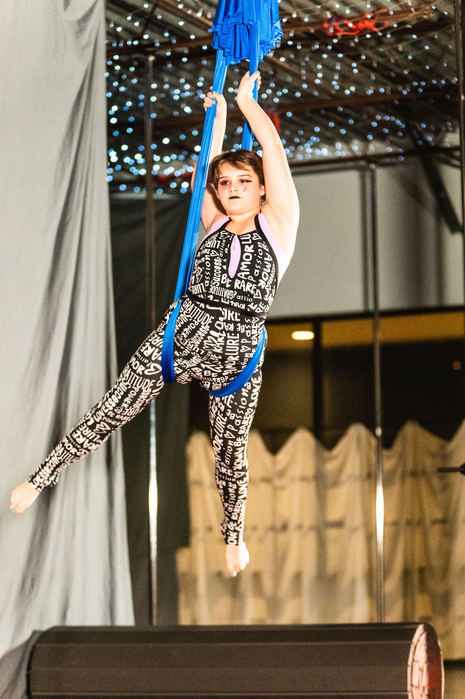Aerial artist in patterned outfit performs split pose while suspended on blue silk fabric.