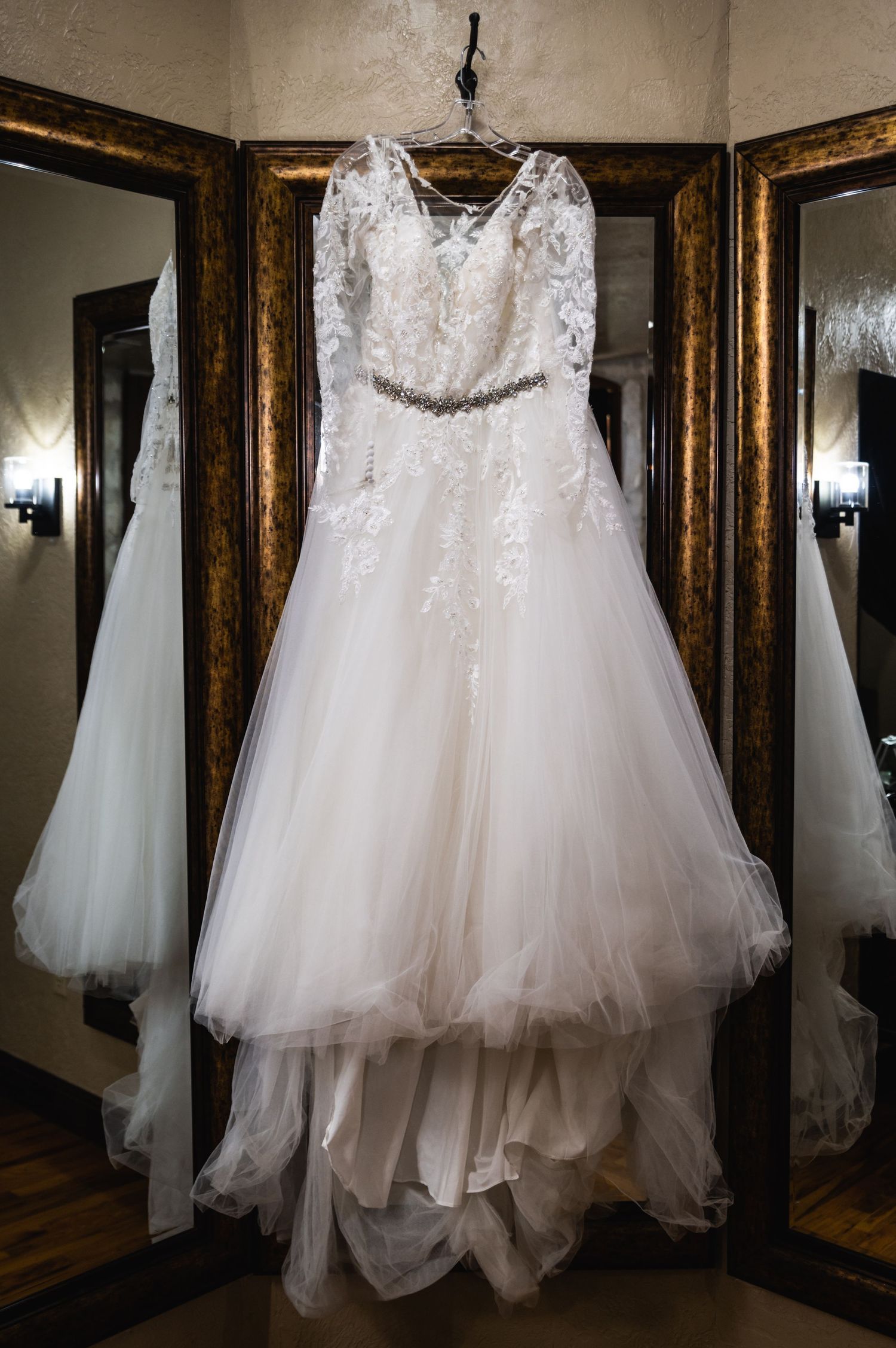 A white lace wedding dress with tulle skirt hangs in an ornate mirror.