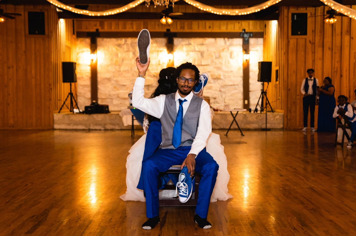 Groom in blue suit celebrating on chair at rustic wedding reception venue with warm lighting.