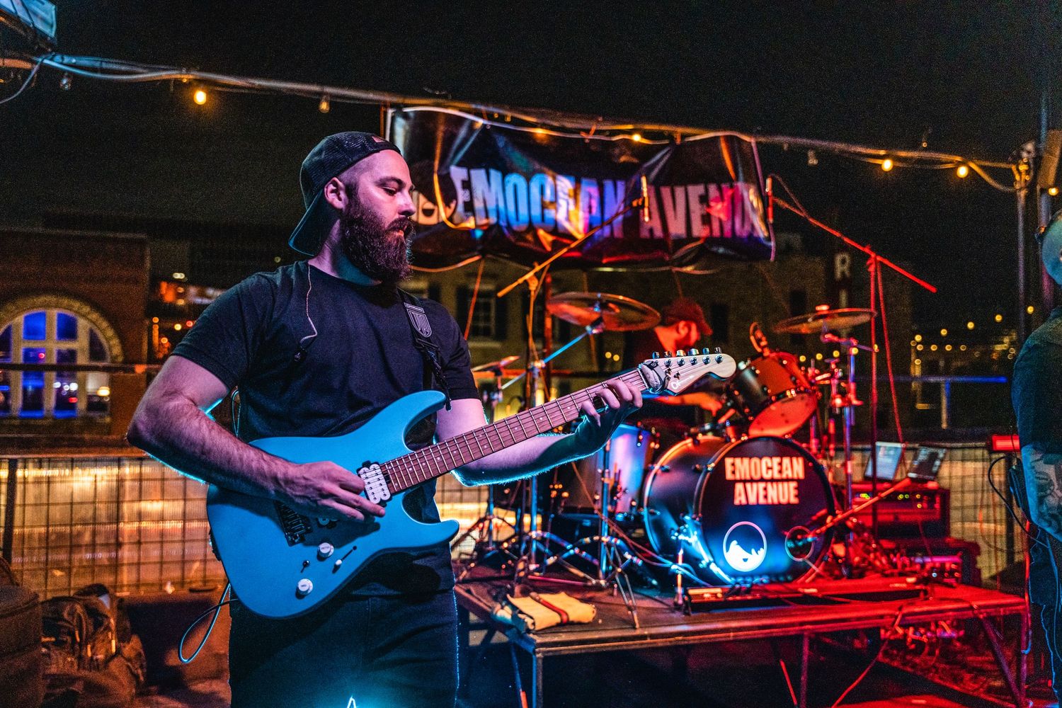 Guitarist plays blue electric guitar under illuminated venue sign at night.