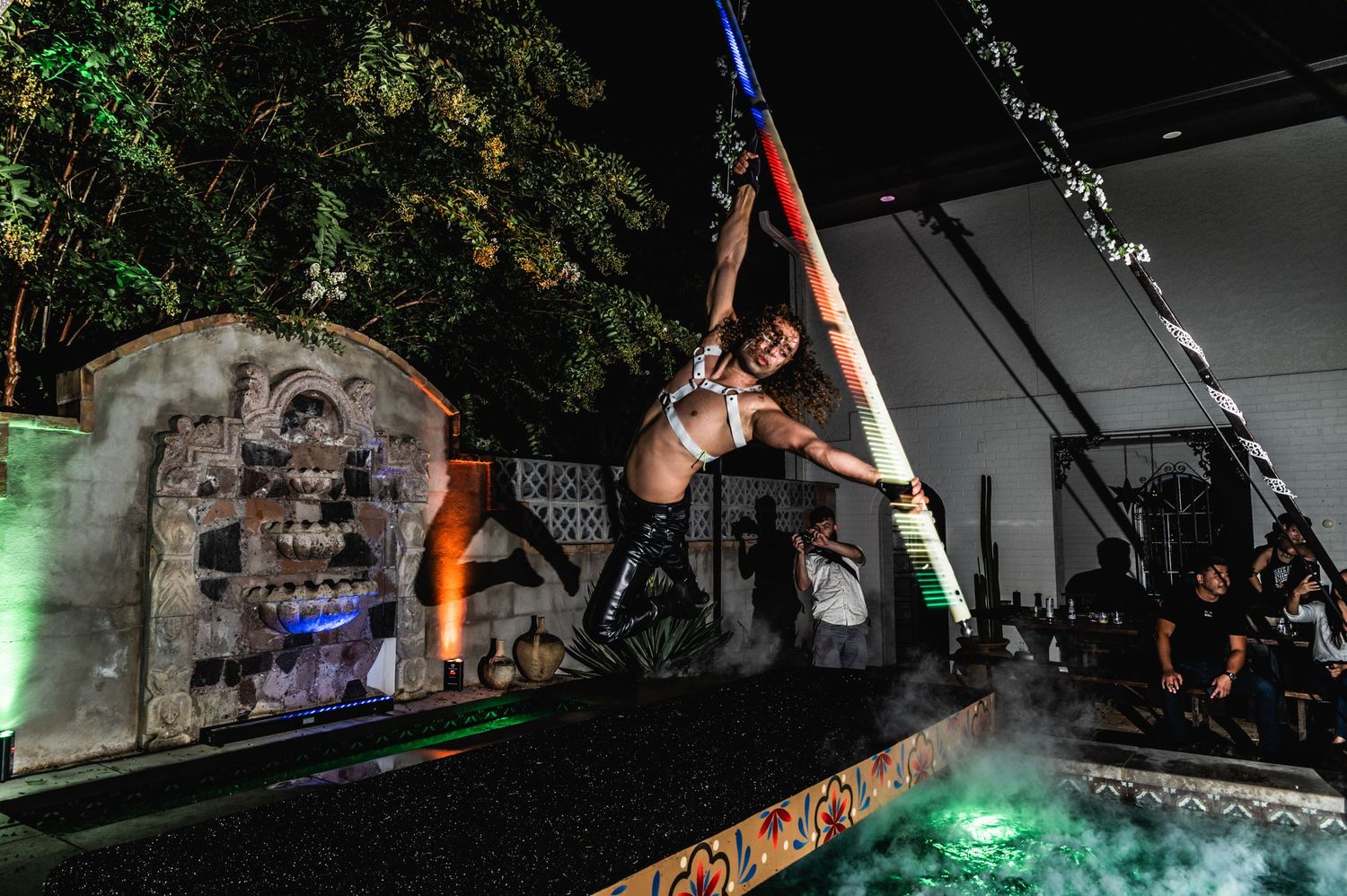 Acrobatic performer swings on aerial equipment against illuminated backdrop during nighttime show.