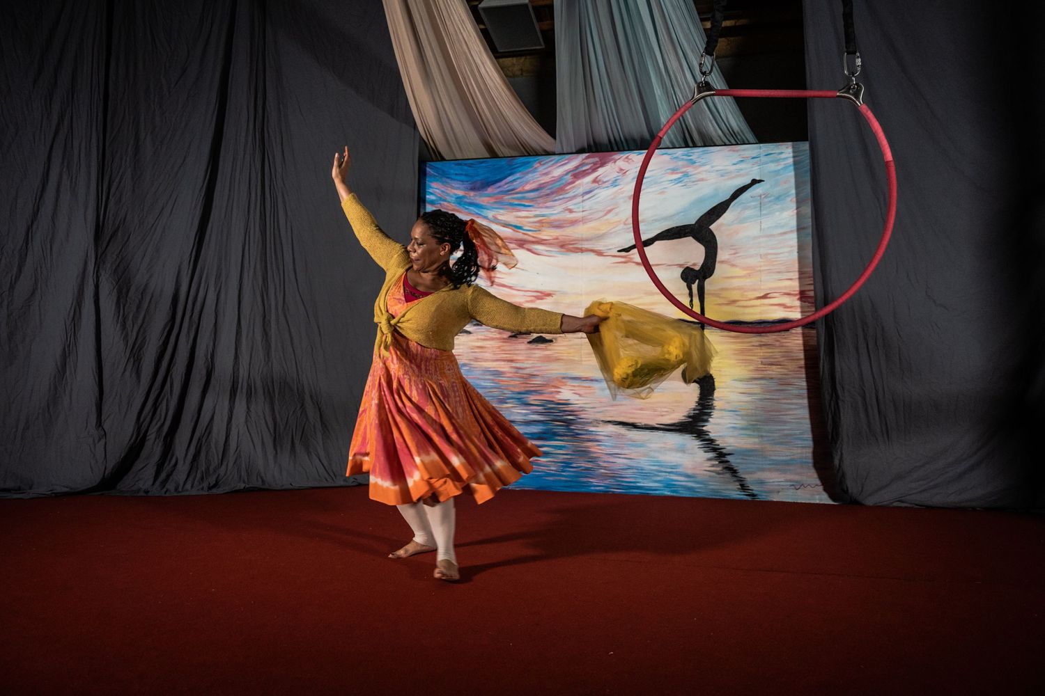 Dancer in flowing orange dress performs gracefully against a painted backdrop on stage.