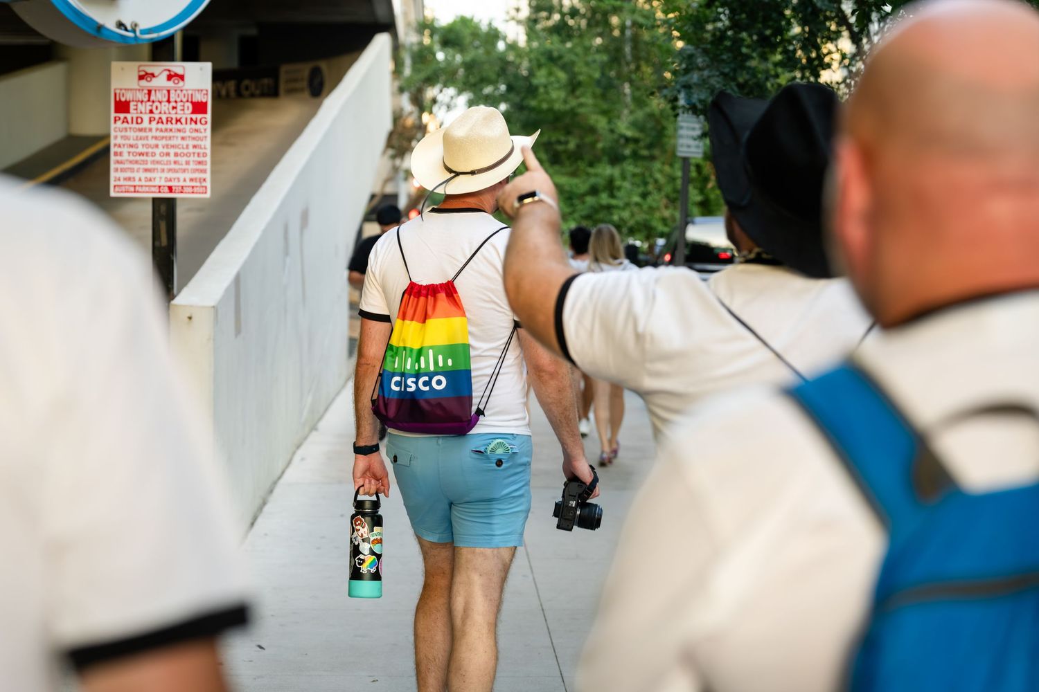 Person wearing rainbow backpack and hat walking on sunny sidewalk at Pride event with camera in hand.