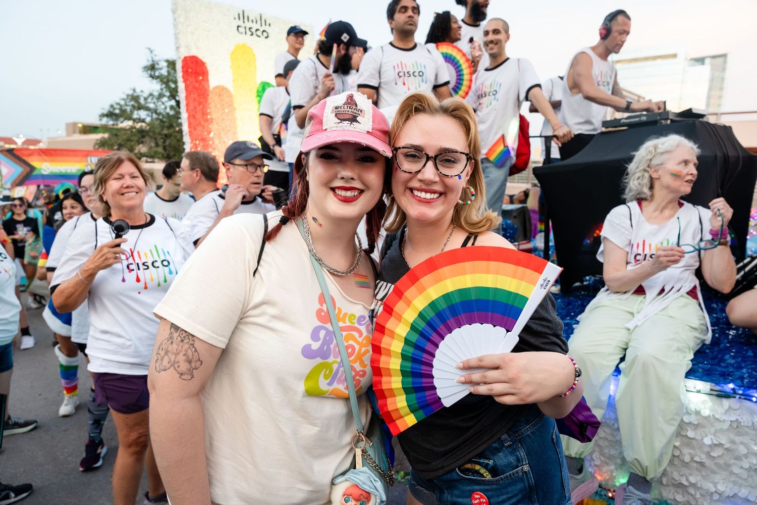 Two women smile at a Pride parade, one holding a rainbow fan, with colorful floats and crowd behind them.