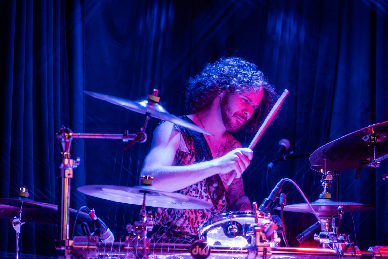 Drummer performs energetically on stage with blue and purple concert lighting creating a dramatic atmosphere.